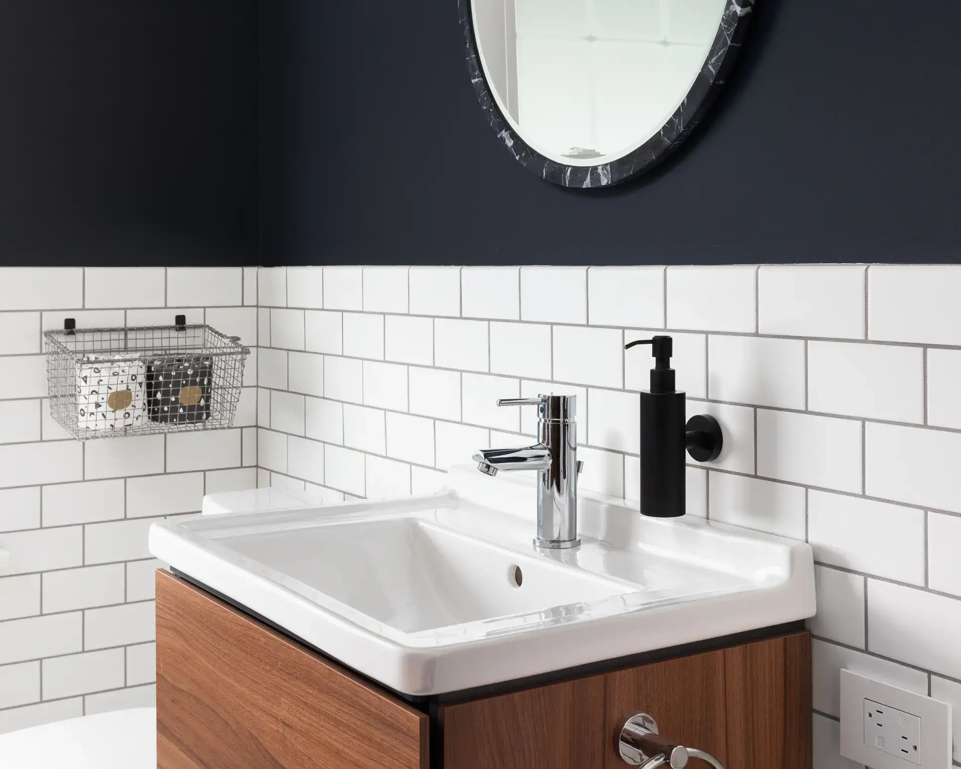 A white basin on a walnut vanity against white subway tile and a navy wall